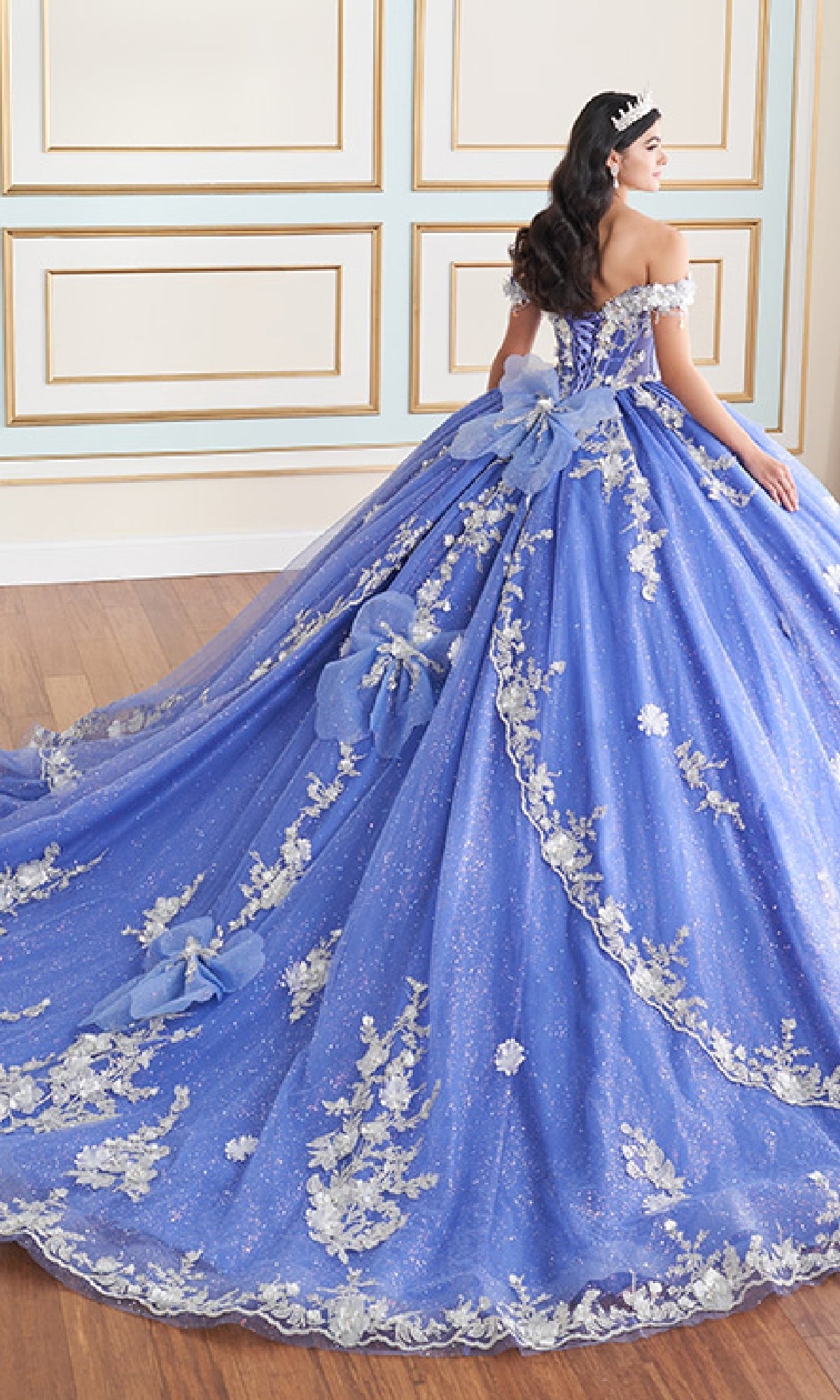 A woman wearing the Quinceanera Dress PR30178 by Princesa, featuring sparkling blue fabric, floral appliqué, large bows, and a corset bodice with tiara, stands in an ornately decorated room with wood floors and paneled walls.